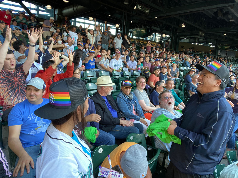 A large group of people sitting in seats at a baseball stadium turn and smile to the camera. A woman in the center wears a t-shirt that says "The Future Takes Transit."