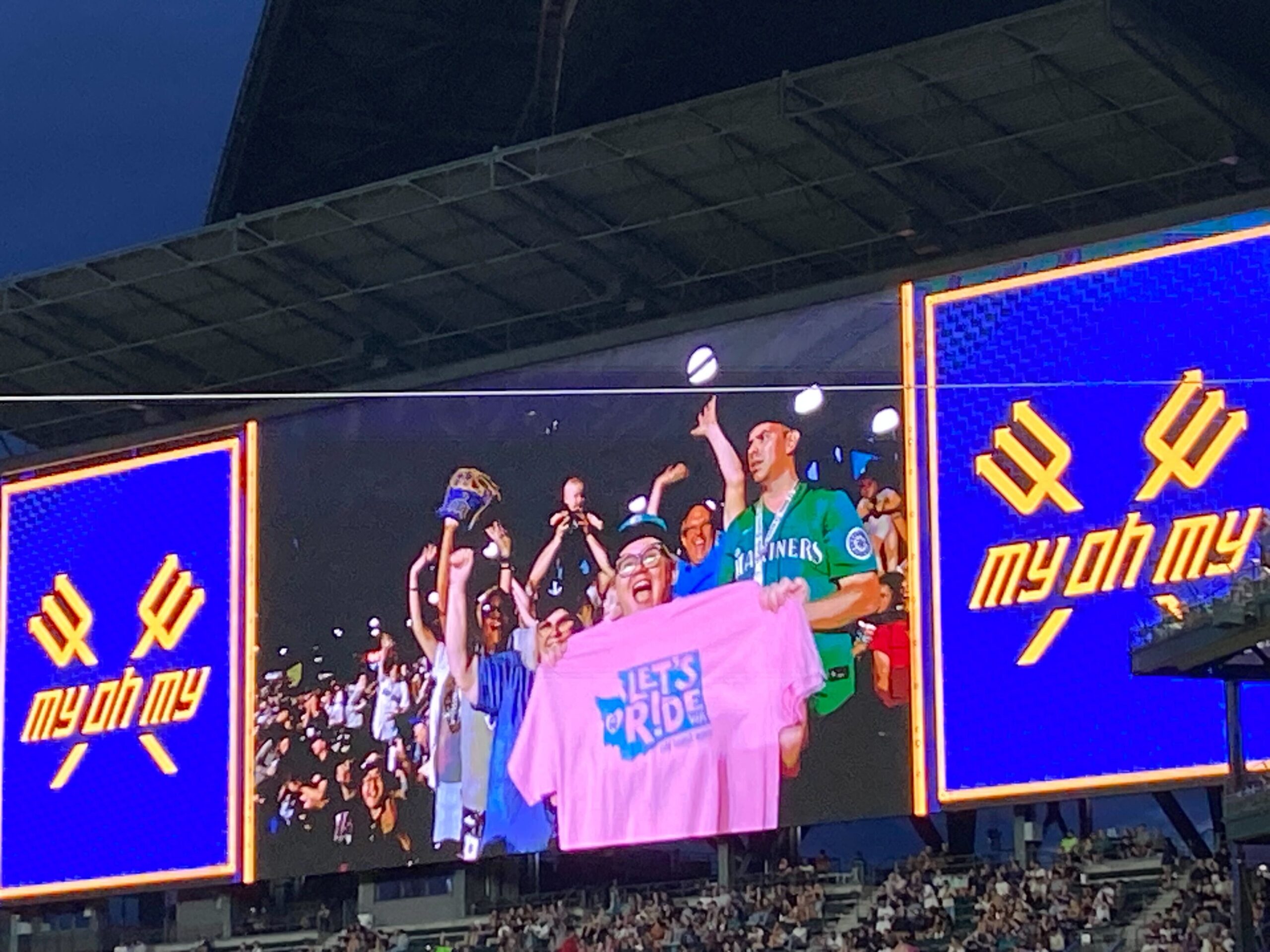 Image of the Jumbotron at a Mariners game. Screen shows a fan holding up a "Let's Ride, WA" pink t-shirt