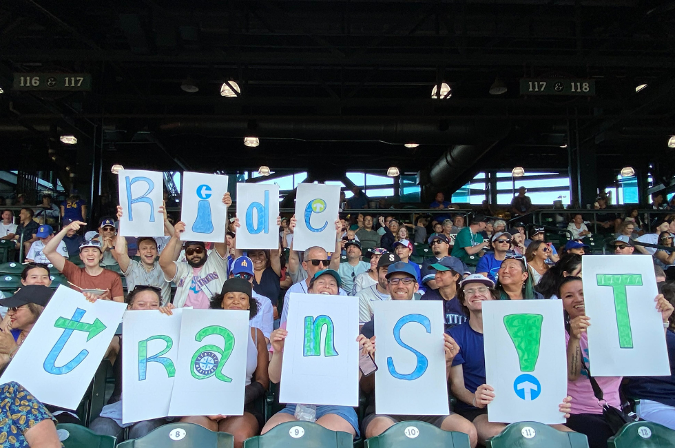 Transit fans at a Mariners game hold up signs that spell out "RIDE TRANSIT!"