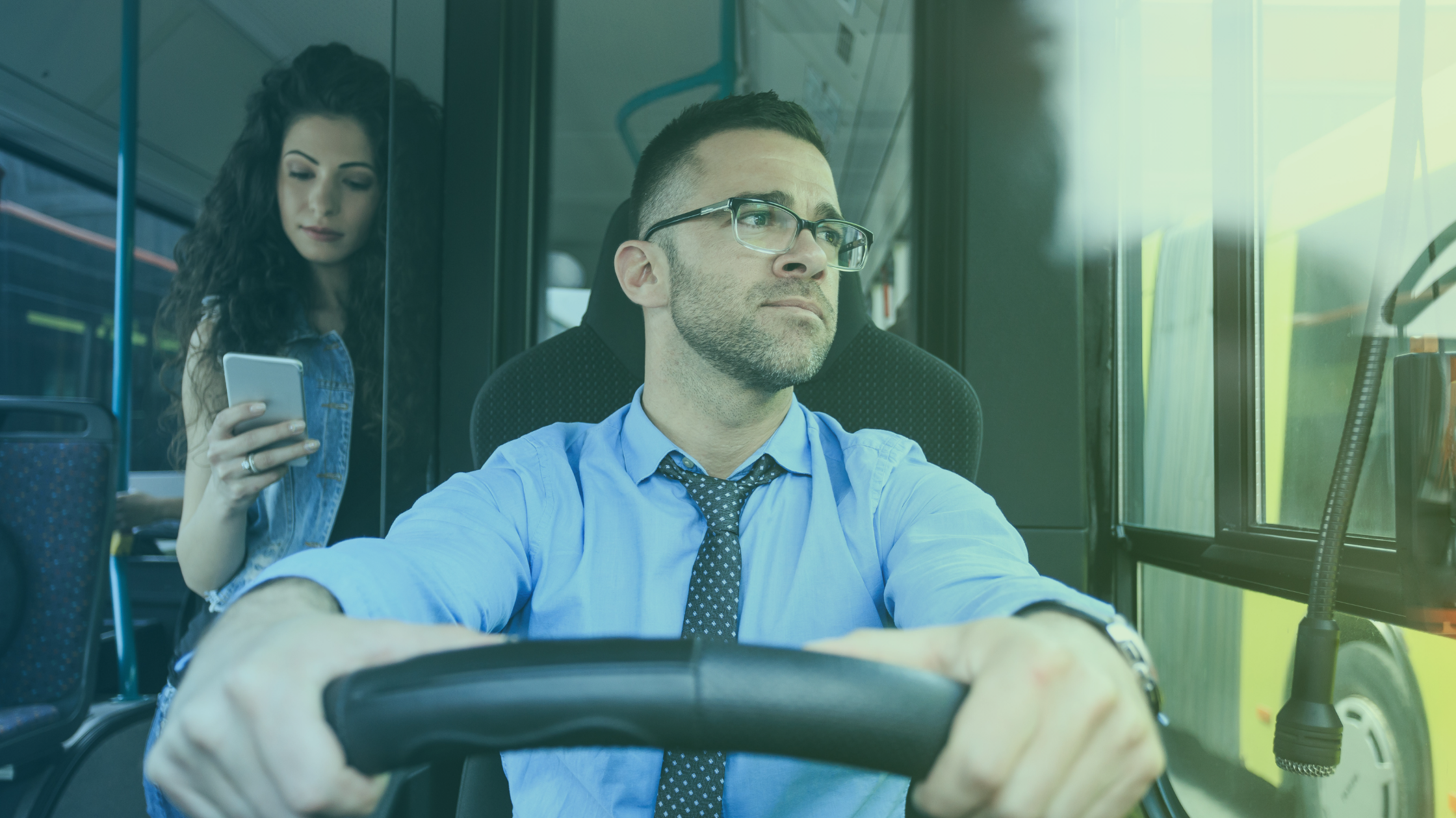 Photo of a bus driver behind the wheel with a woman on her phone in the background. 