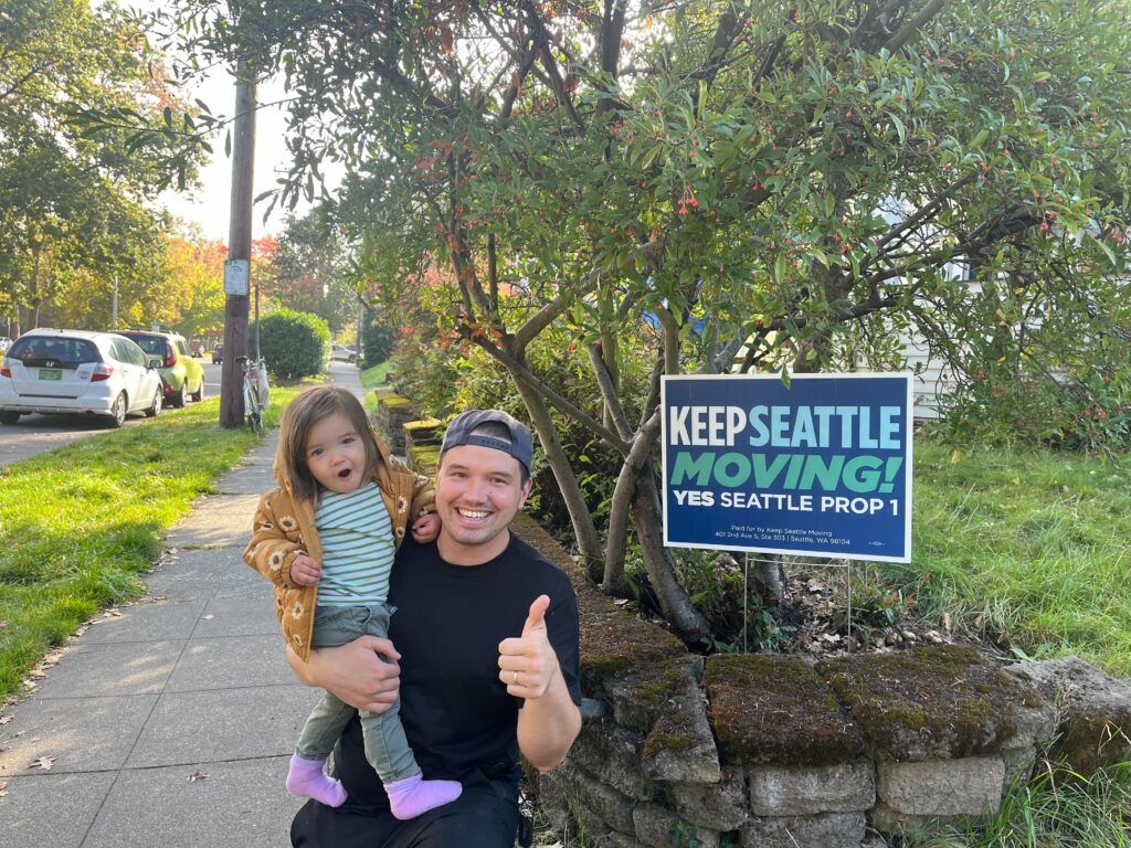 A dad poses with his daughter with a thumbs up and a KEEP SEATTLE MOVING sign in the background. 