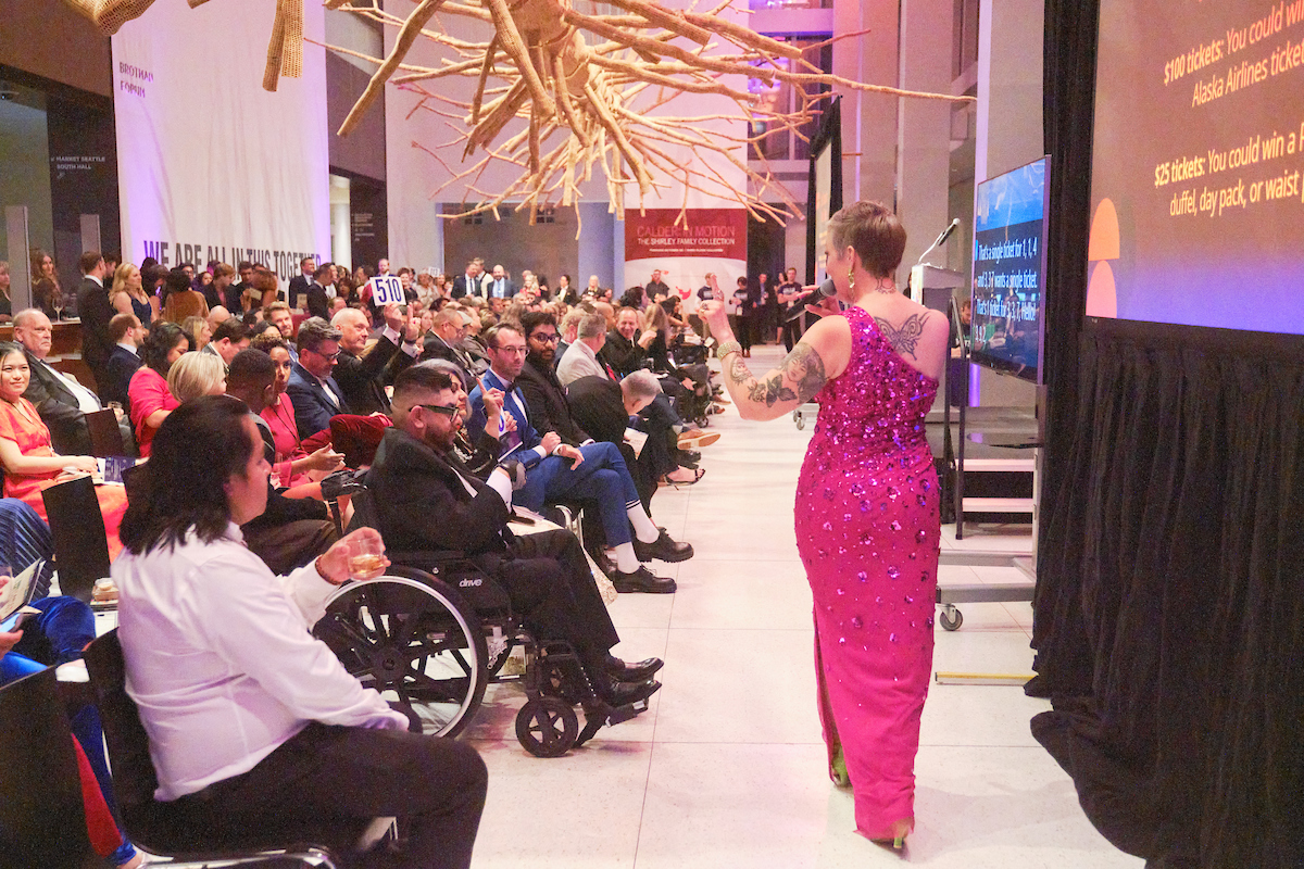 Emcee in a pink dress paces before a crowd of attendees at a gala.