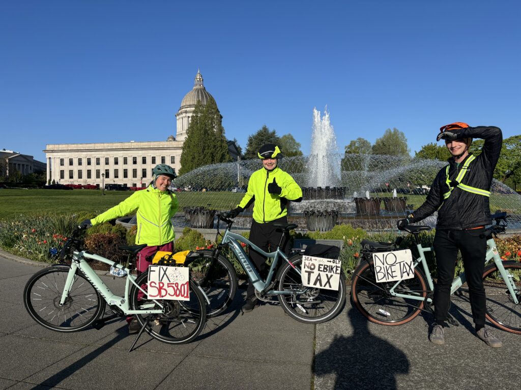Three bikers pose in front of the capitol building in Olympia, WA with e-bikes and signs that say