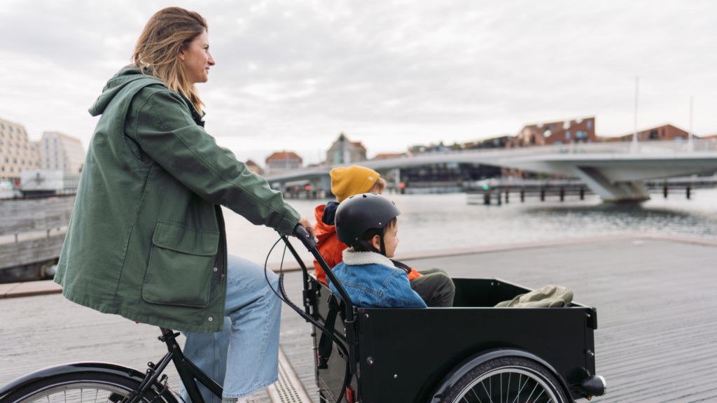 A woman bikes along a riverfront boardwalk with two children in a cargo bucket in front of her. 