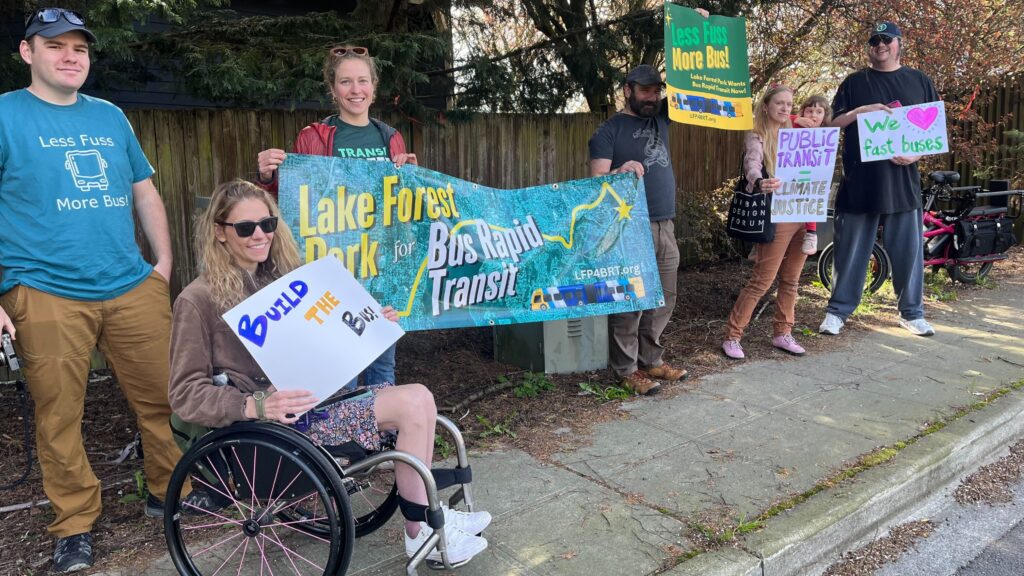 People gather on a sidewalk next to SR-522 holding signs that say things like 