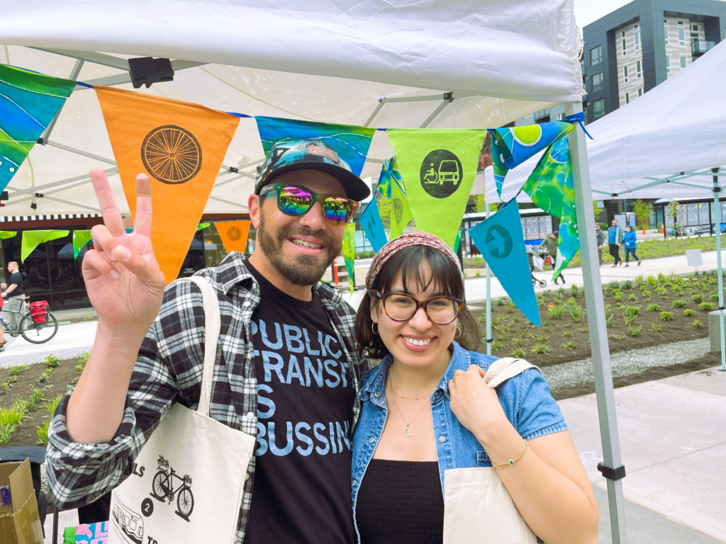 A couple stand in front of brightly colored flags -- the man holds up a peace sign.