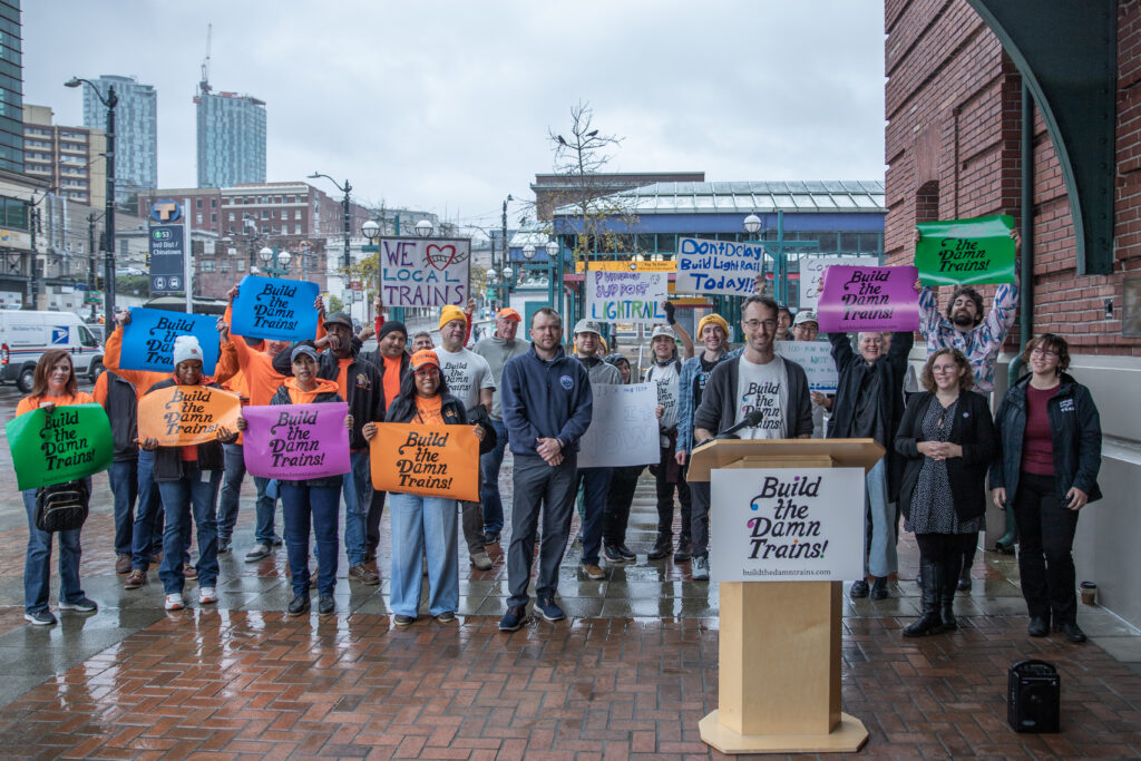 Transit advocates pose in front of the International District Station with colorful signs that say 