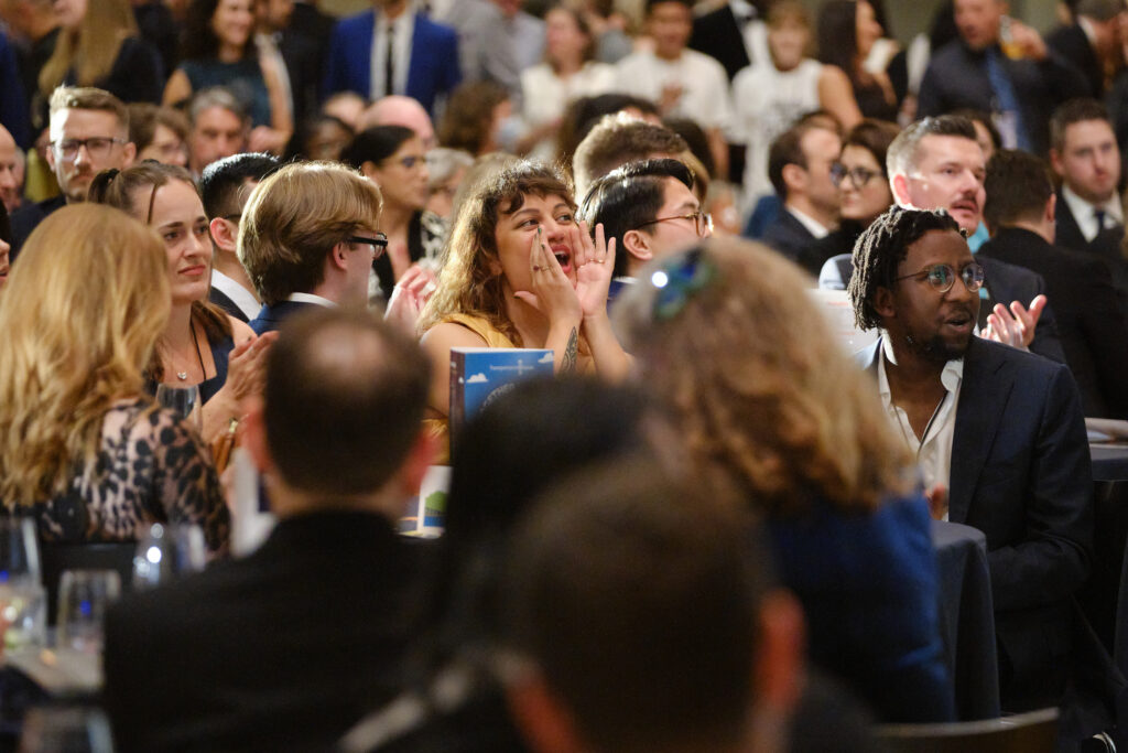 A crowd of people watch the stage. One of them holds their hands to their mouth to give out a whoop!