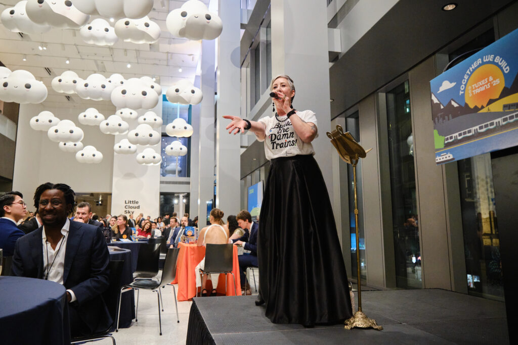 Emcee and auctioneer Sasha Summer Cousineau gestures at the crowd. She's wearing a Build the Damn Trains t-shirt and an art installation of clouds is visible above guests seated at tables. 