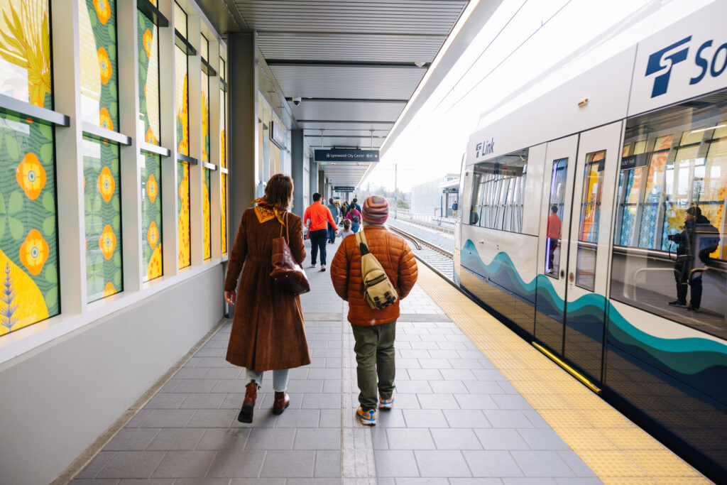 Transit riders in fall colors walk beside a Link light rail train at the platform in Star Lake.