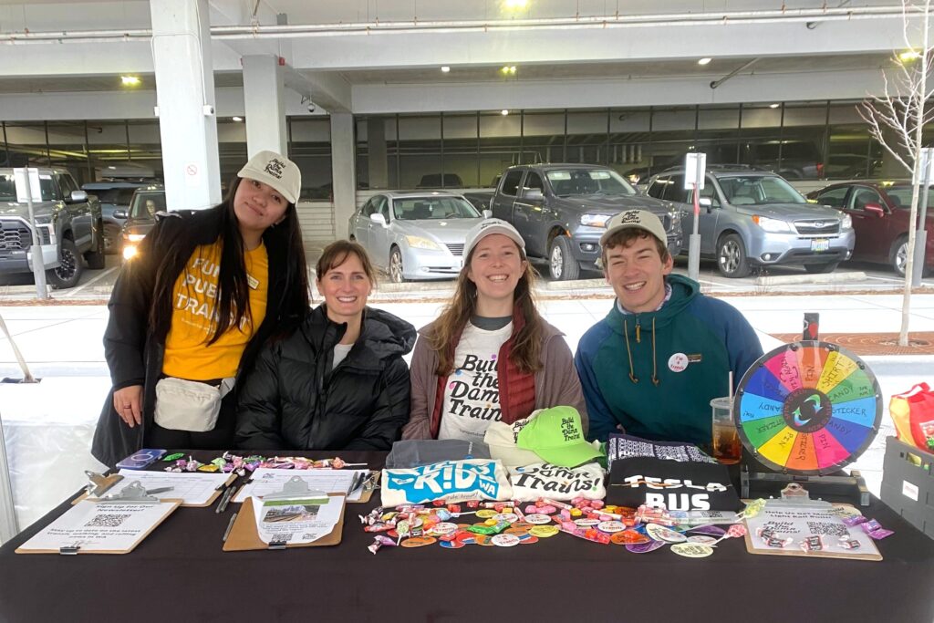 Four TCC staff members sit behind a table covered in colorful stickers, t-shirts, and a prize wheel.