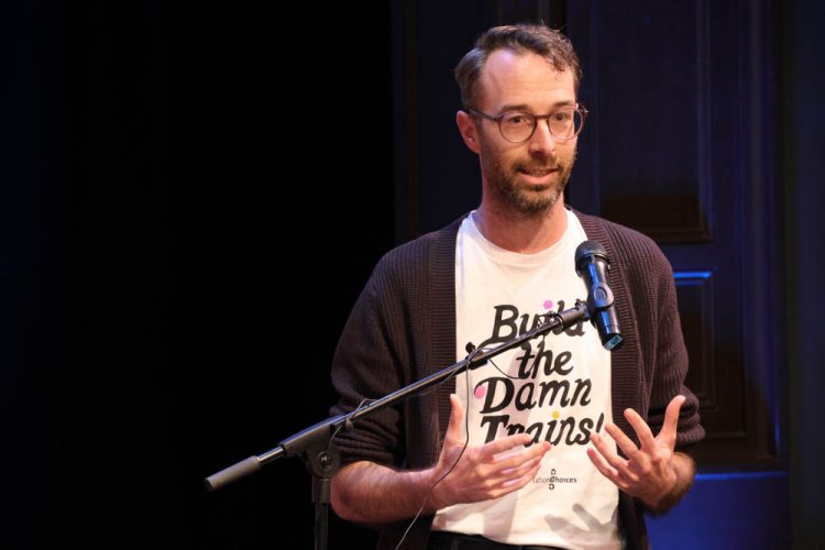 Kirk Hovenkotter speaks before a microphone wearing a "Build the Damn Trains" t-shirt