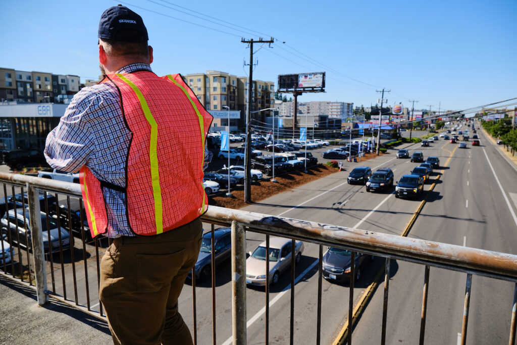 A man walks through the foreground on an overpass. In the background, cars line up on a 6 lane street.