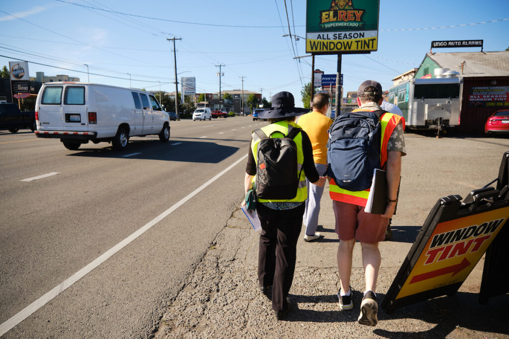 Three people with safety vests are walking away from the camera in the foreground, on the shoulder of a street. A white car passes them on the left.