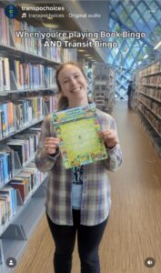 A woman in a library smiles, holding up a Book Bingo card. Text: "When you're playing Book Bingo AND Transit Bingo"
