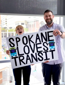 Two smiling people (Erik Lowe and Sarah Rose of Spokane Reimagined) hold up a sign that says "Spokane loves transit" 
